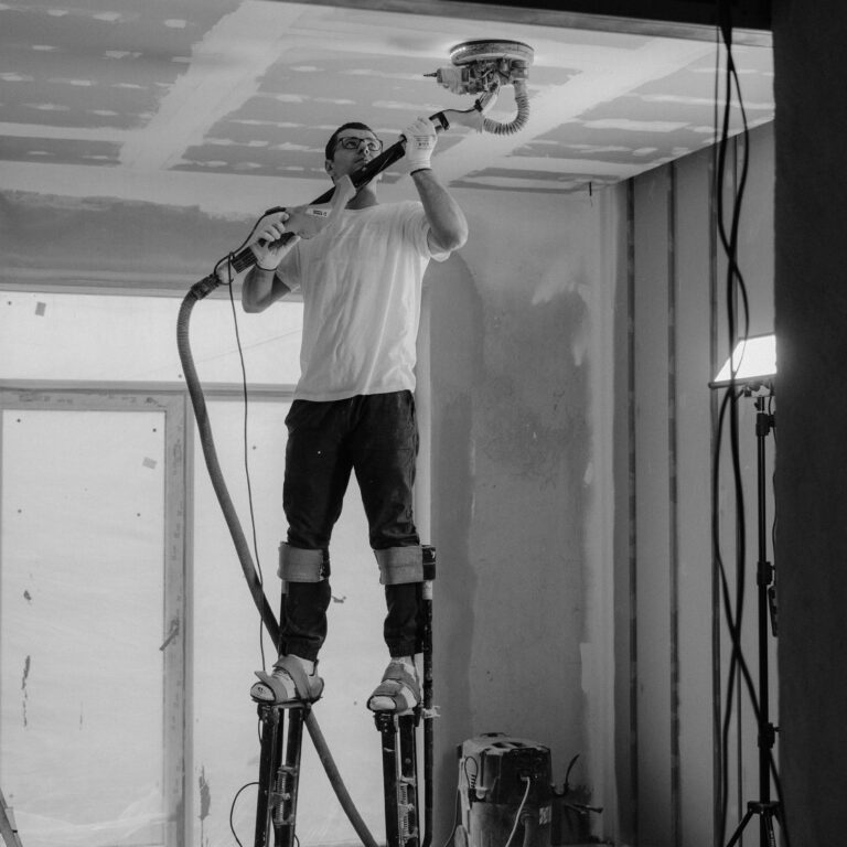 Worker using stilts and power sander on ceiling in construction site.